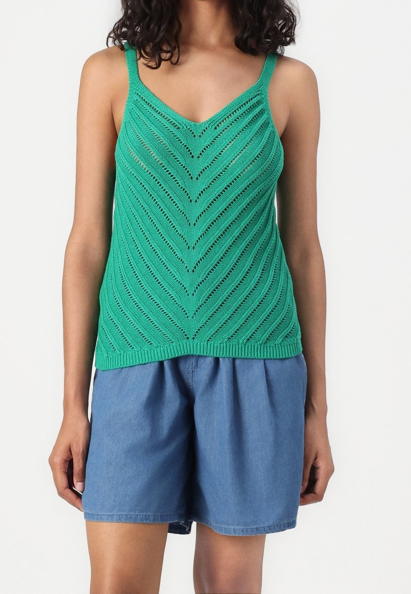 Femme portant un haut en maille sans manches vert à motifs chevrons et un short bleu ample, debout devant un fond blanc.
