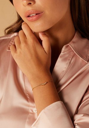 Gold bracelet with a fine chain, featuring three small clear gemstones. The hand rests against a soft pink satin shirt.