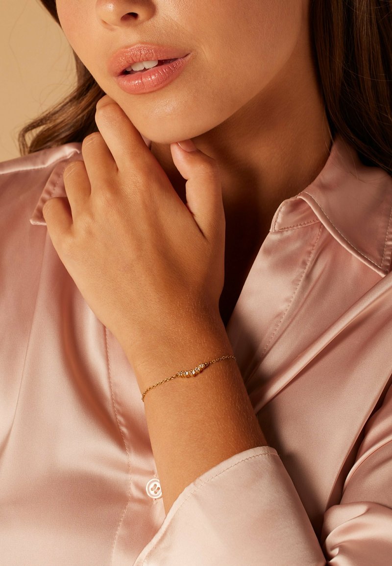 Gouden armband met een fijne ketting, voorzien van drie kleine heldere edelstenen. De hand steunt tegen een zachtroze satijnen shirt.