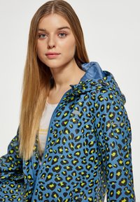 Mėlynas lietpaltis su juodu ir citrinos žaliais leopardų raštais, su kapišonu, užtrauktuku ir sagų akcentais. Glotnus, blizgus audinys.