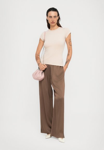 Donna con tatuaggi che indossa una maglietta beige a maniche corte, pantaloni larghi marroni, scarpe bianche a punta e porta una piccola borsa rosa con tracolla a catena.