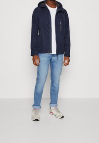 Navy lightweight jacket with a hood, zippered chest pocket, paired with light blue rolled-cuff jeans and grey sneakers with pink accents.