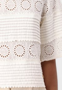 Witte blouse met geplooide stof en kant met zon- en sterpatronen. Golvende randen accentueren het ontwerp, wat zorgt voor textuur en detail.