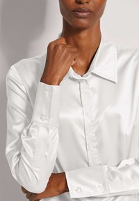 Persoon in een wit satijnen overhemd met knopen, hand rustend bij de kin, gecropt bij de schouders en boven de taille.