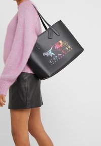 Sort lædertaske med grafisk design, der viser en vogn, dyr og logo. Tasken har to skulderremme og en tekstureret overflade.