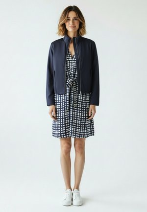 Frau steht, trägt eine marineblaue Jacke über einem marineblauen, gemusterten Kleid mit gebundener Taille und weißen Turnschuhen, vor einem schlichten hellen Hintergrund.