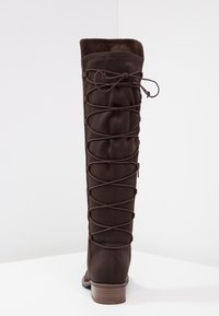 Hnědá kožená vyšší bota vyrobená z měkkého materiálu, s krajkovým designem na zadní straně, kulatou špičkou a nízkým klínovým podpatkem.