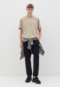 T-shirt in cotone beige, pantaloni cargo neri, camicia a quadri bianca e nera annodata in vita, sneakers nere, sfondo bianco.