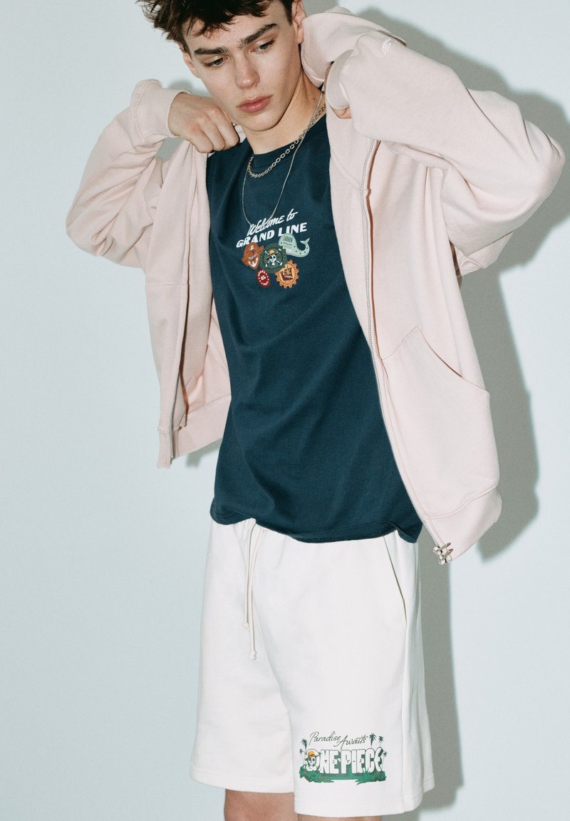 Jeune homme ajustant un sweat à capuche zippé rose clair, portant un t-shirt graphique bleu marine avec patchs et un short blanc avec le logo "One Piece" et des palmiers.