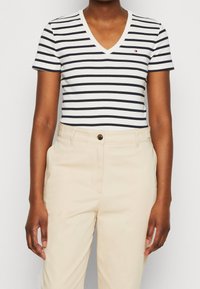 Tričko s v-neck výstřihem a krátkým rukávem v námořnické modré a bílé horizontální pruhy; spárováno se světle béžovými kalhotami s předním zapínáním na knoflík.