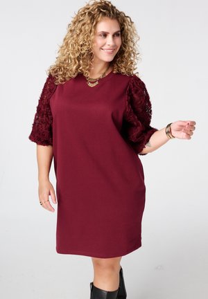 Bordeaux jurk met korte, getextureerde mouwen versierd met bloemmotieven. Voorzien van een ronde halslijn en een ontspannen pasvorm. Gepaard met zwarte laarzen.