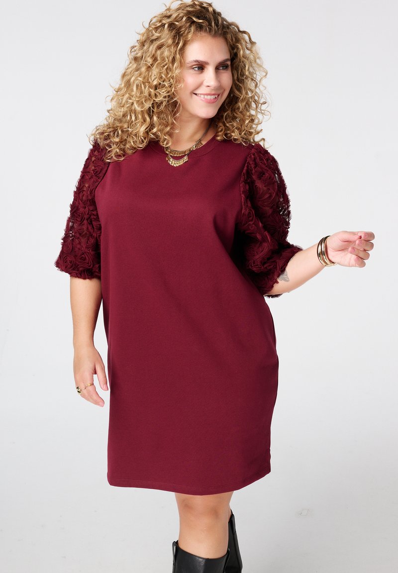 Robe bordeaux avec des manches courtes texturées ornées de motifs floraux. Caractéristiques d'un col rond et d'une coupe décontractée. Associée à des bottes noires.