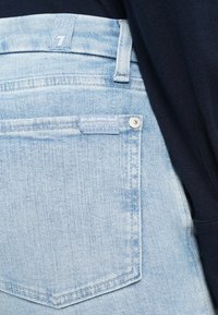 Poche de jean en denim bleu clair avec une petite étiquette en tissu et un rivet en métal, partiellement recouverte par un tissu bleu marine foncé.
