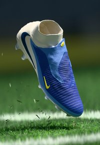 Chuteira de futebol azul com um upper texturizado, colarinho branco, detalhes amarelos e um design aerodinâmico, pairando sobre a relva com detritos.