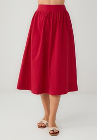 Jupe rouge, longueur genou avec une taille élastique, présentant un design froncé et une texture en coton lisse. Portée avec des sandales beiges.