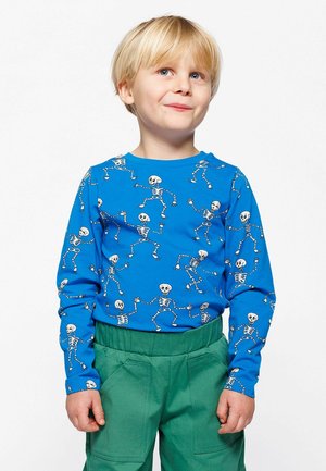 Blaues Langarmshirt mit einem weißen Skelettdruck. Material ist glatte Baumwolle. Kombiniert mit grünen Hosen mit strukturiertem Finish.