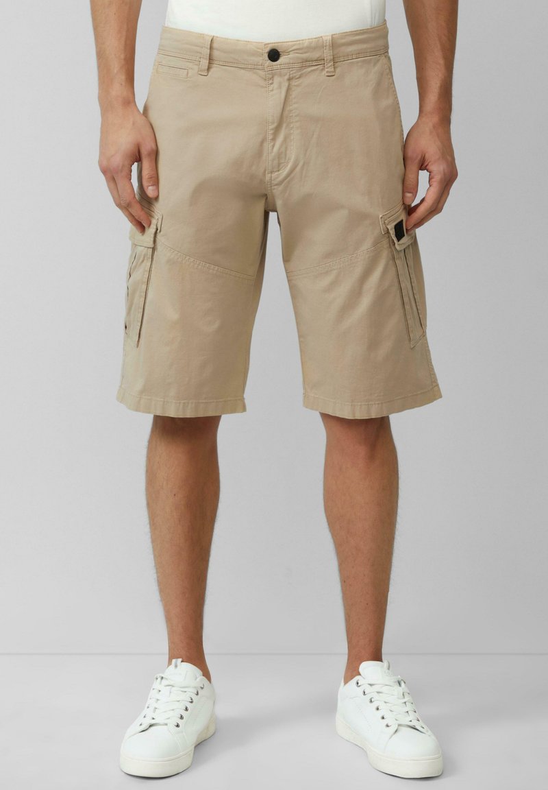 Shorts cargo beiges avec une coupe décontractée, en tissu léger. Dotés de poches appliquées, de passants de ceinture et d'ourlets roulés. Associés à des baskets blanches.