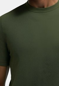 T-shirt a maniche corte, con collo rotondo, di colore verde oliva scuro. Presenta un tessuto testurizzato a coste con sottili linee diagonali. Finitura liscia.
