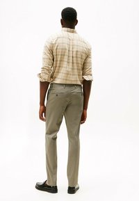 Beige geruite overhemdd met opgerolde mouwen; grijze broeken, rechte pasvorm; zwarte dresschoenen met een gladde afwerking. Gezicht vanaf de achterkant.