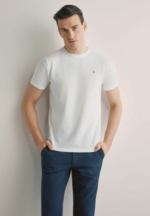 Młody mężczyzna o ciemnych włosach, ubrany w gładką białą koszulkę z krótkim rękawem i ciemnoniebieskie spodnie, stoi z rękami w kieszeniach na tle jasnej ściany.