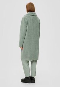 Manteau en polaire vert clair texturé avec col châle, manches longues et coupe mi-longue, associé à un pantalon vert clair et des bottes noires.
