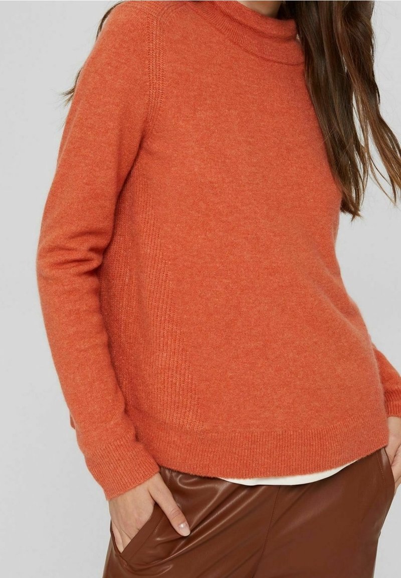 Oranssi neulepaita, jossa on korkea kaulus, ribatut hihansuut ja helma. Paitaa käytetään ruskeiden tekonahkahousujen päällä, joissa on sileä pinta ja rento istuvuus.