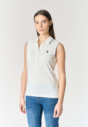 Donna che indossa una polo bianca senza maniche con un piccolo logo nero sul petto, abbinata a jeans blu, in piedi davanti a uno sfondo chiaro uniforme.