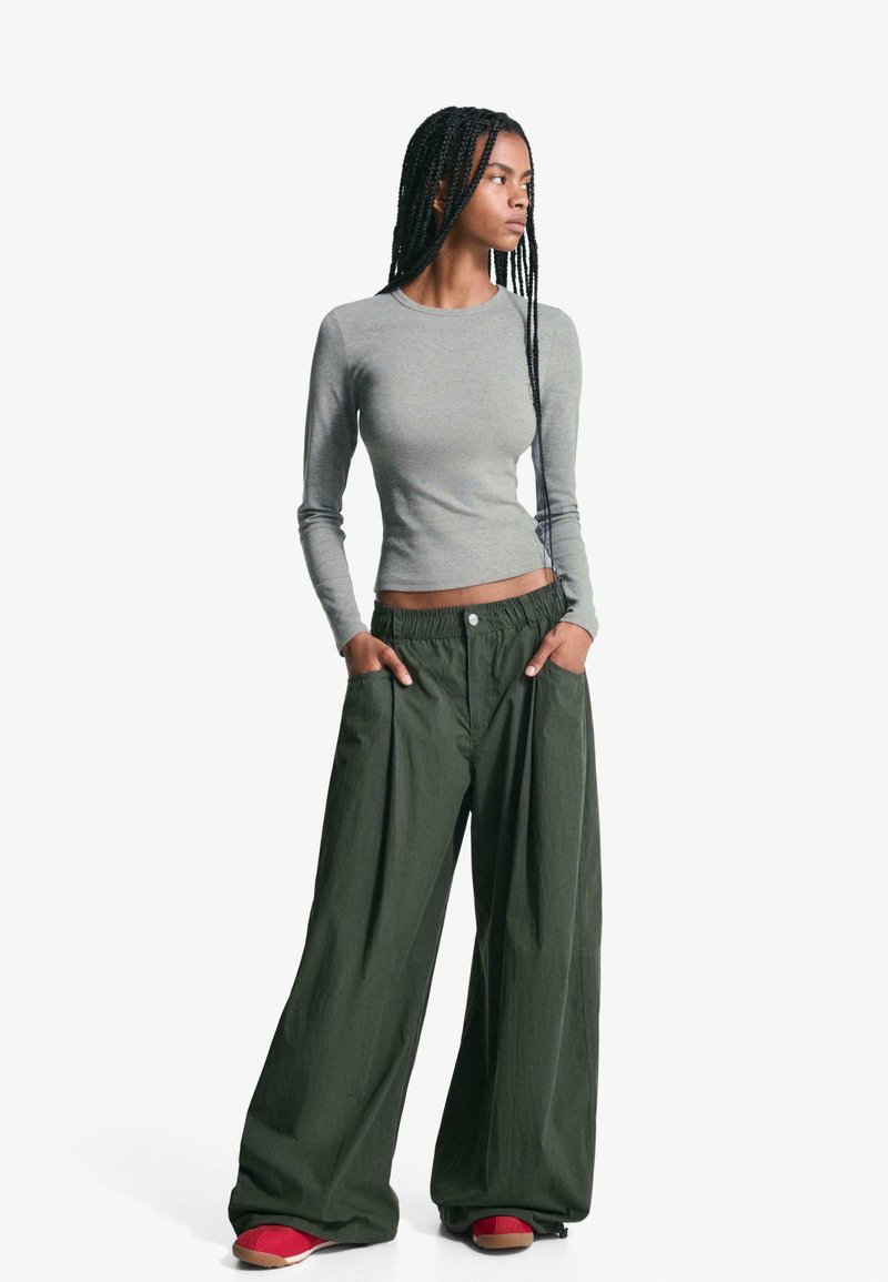 Top gri cu mâneci lungi, strâmt, combinat cu pantaloni verzi largi, comozi, cu talie elastică și buzunare, purtați cu pantofi roșii.