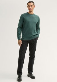 Grøn strikket sweater med rund hals, båret med sorte bukser og sorte snørebåndsstøvler. Glat tekstur, afslappet pasform, minimal detaljering.