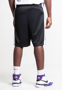 Zwarte mesh sportshorts met witte zijstreep, elastische tailleband en een losse pasvorm, gecombineerd met witte en paarse basketbalschoenen.