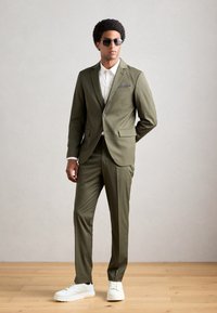 Bărbat îmbrăcat într-un costum verde măsliniu, cămașă albă, adidași albi și ochelari de soare negri, stând pe un podea din lemn, în fața unui perete gri simplu.