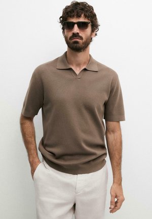 Uomo con capelli ricci e barba che indossa un polo marrone a maniche corte in maglia e pantaloni chiari, in piedi con una mano in tasca, occhiali da sole indossati.