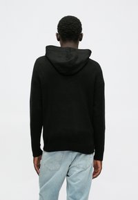 Black hooded sweater with a soft texture, relaxed fit, and ribbed hem, paired with light blue denim jeans. View from the back.