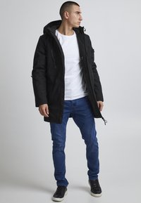 Parka impermeabilă neagră cu glugă, buzunare cu fermoar și șireturi ajustabile. Asortată cu un tricou alb și blugi blau.
