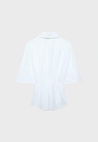 Blouse blanche à manches courtes avec col large et plis dans le dos, présentée à plat sur un fond gris clair.