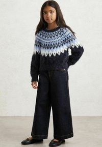 Kék és fehér mintás bolyhos pulóver, kerek nyakkal, széles szárú sötét farmernadrággal és fekete lapos cipőkkel párosítva, semleges háttér előtt.