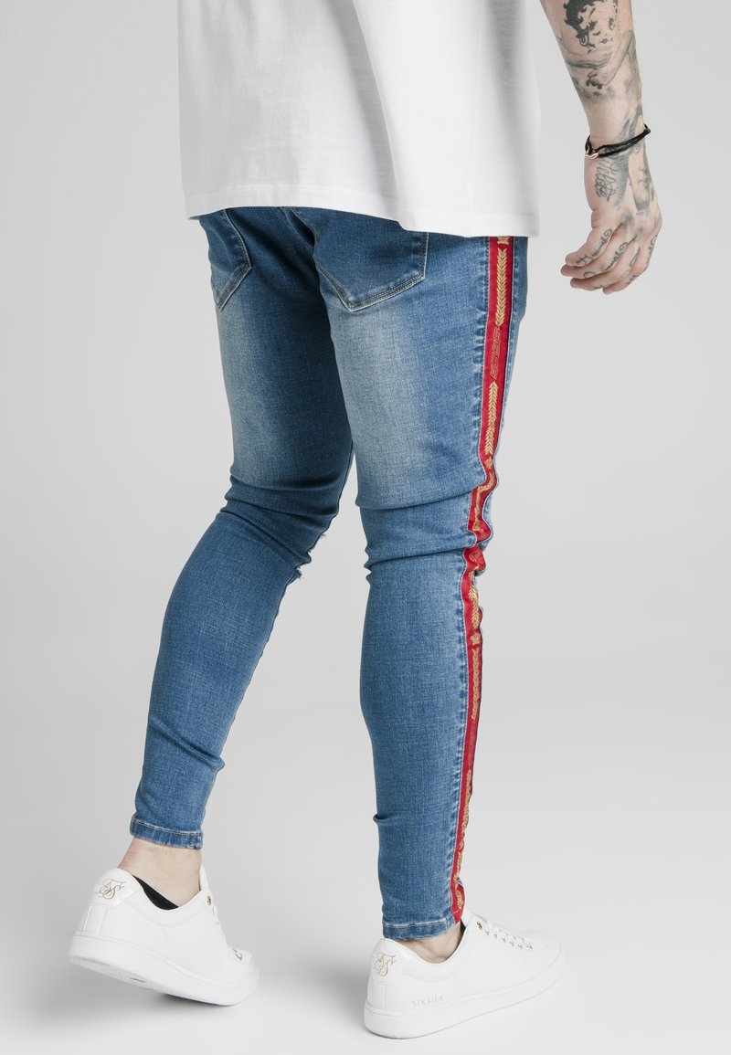 Tunna blå denimjeans med röda och gula sidostråkar, med en lätt blekning. Bärs med vita sneakers, vilket framhäver en avslappnad stil.