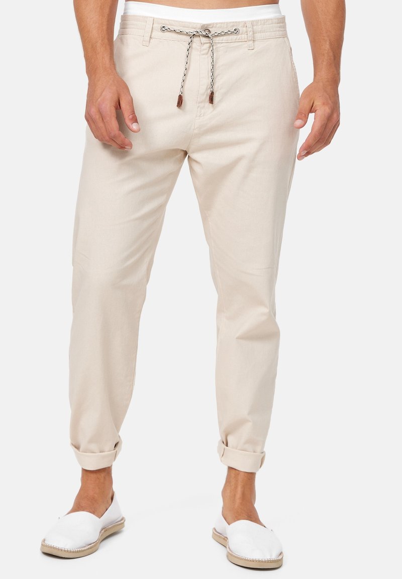 Homme portant un pantalon beige retroussé avec un cordon noir et blanc et des chaussures blanches sans lacets, debout devant un fond uni.