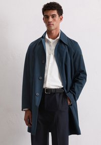Marineblauer Trenchcoat mit glatter Textur, breitem Kragen und drei großen Knöpfen. Kombiniert mit einem weißen Hemd und einem schwarzen Gürtel, der ein elegantes Design bietet.