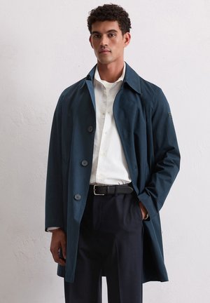 Marineblauwe trenchcoat met een gladde textuur, brede kraag en drie grote knopen. Gecombineerd met een wit shirt en een zwarte riem, met een strak ontwerp.