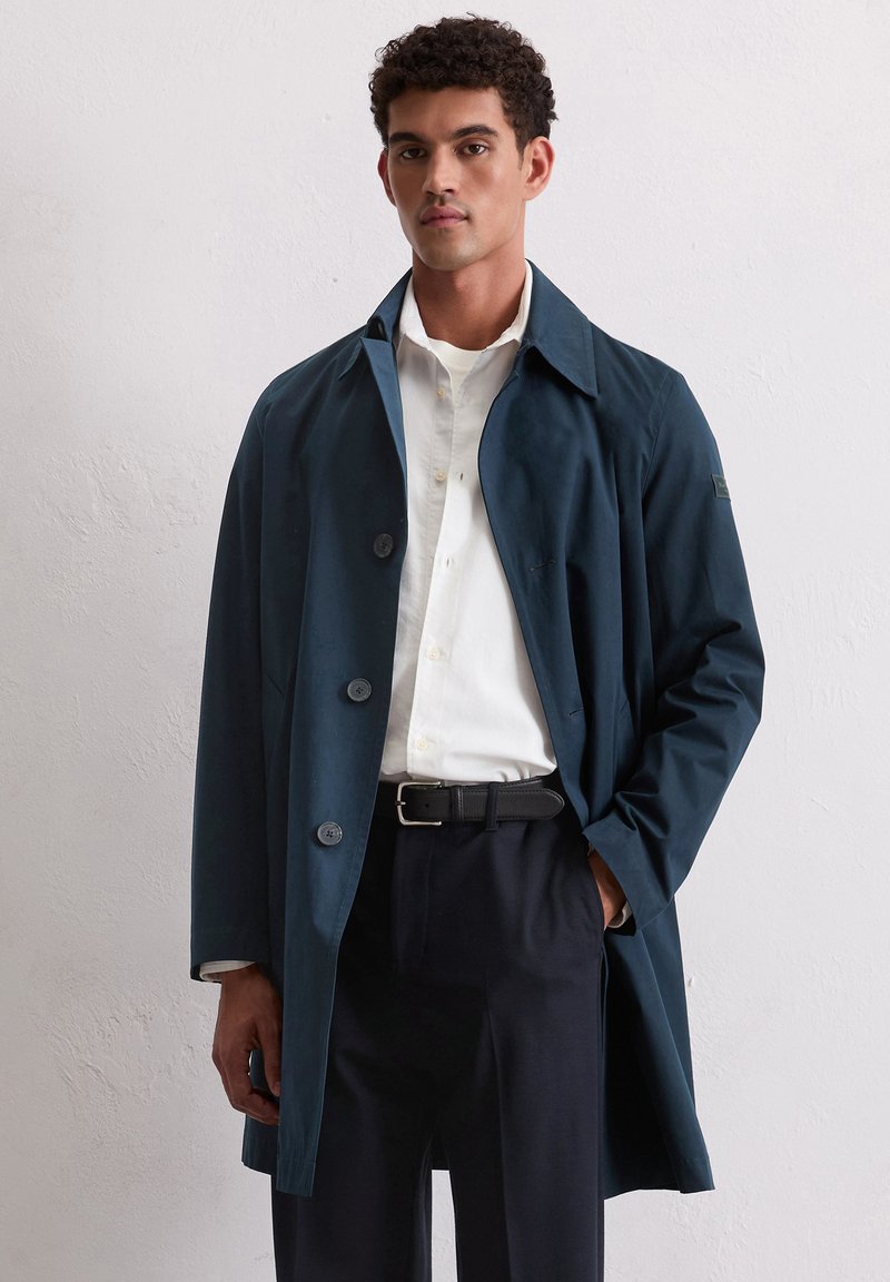 Marineblauer Trenchcoat mit glatter Textur, breitem Kragen und drei großen Knöpfen. Kombiniert mit einem weißen Hemd und einem schwarzen Gürtel, der ein elegantes Design bietet.