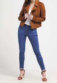 Bruine suède jack met een cropdesign, gecombineerd met blauwe jeans en open teen hakken. Het jack heeft ritsaccenten en een kraag.