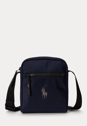 Pequeño bolso bandolera azul marino con correa negra ajustable, bolsillo frontal con cremallera y logotipo bordado de jugador de polo en el centro.