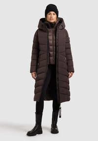 Brun pufferjakke med hætte, vertikal quiltet design og lynlås, lagd over en kortere jakke. Sorte leggings og støvler fuldender looket.