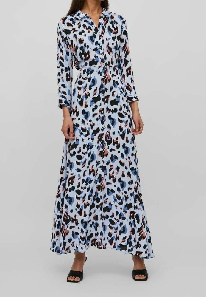 Femme portant une robe maxi à manches longues avec col, imprimé léopard abstrait bleu, noir et marron, et des talons ouverts noirs.