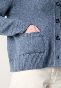 Personne portant un cardigan en tricot bleu avec des boutons, main partiellement dans la poche avant, associé à un pantalon blanc.