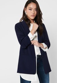 Blazer bleu marine avec un design ajusté, revers crantés et poches avant, porté sur une chemise blanche à boutons, associé à un jean foncé.