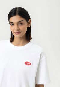 Witte katoenen t-shirt met een geborduurde rode lippen graphic op de linker borst. Klassieke pasvorm met een ronde hals en korte mouwen.