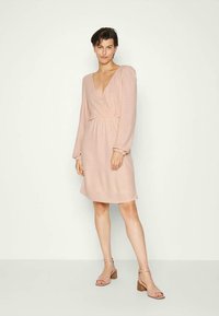 Vestido envolvente rosa claro com mangas longas, tecido texturizado, decote em V profundo, cintura ajustada e comprimento até o joelho. Com sandálias de salto leves.