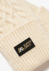 Bonnet tricoté couleur crème avec des motifs torsadés et côtelés ; comporte une étiquette en tissu noire avec du texte doré et un logo.